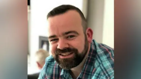 Family Handout Man with dark hair and beard, with a flannel shirt on. 