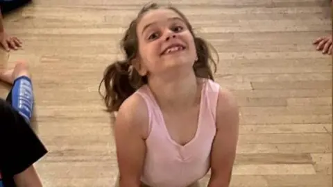 Handout Jessica smiling at the camera. Her brown hair is tied in two ponytails. She is wearing a pink dress.