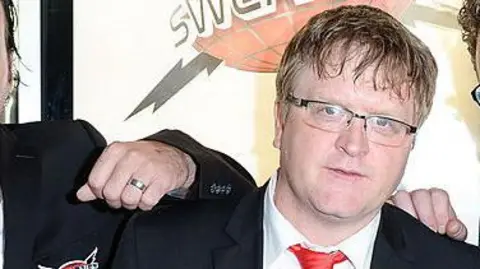 Mike Smith appearing at a film premiere is wearing a dark suit coat and red tie. The hands of two co-stars are seen on his shoulder though they are both out of frame. 