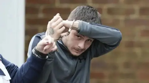 Kyle Dumble in handcuffs. He is attempting to cover his face. He is wearing a grey tracksuit top. 