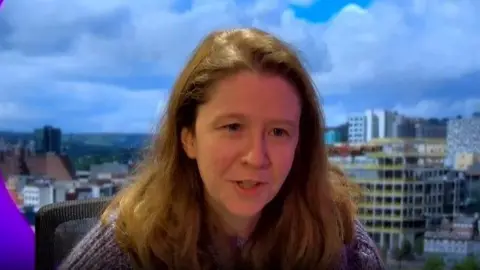 Ms Ellis sits in a studio with an image of Sheffield's skyline in the background. She has long brown hair and is wearing a knitted top.