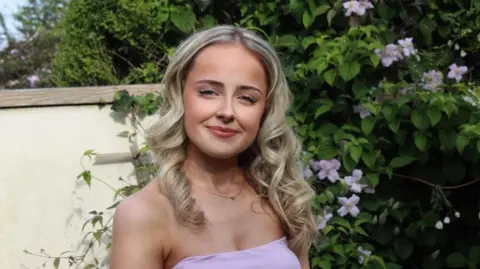 Caitlin Giffard Caitlin, a young woman with wavy blonde shoulder-length hair wearing a pink dress in front of a bush with pink flowers on it.