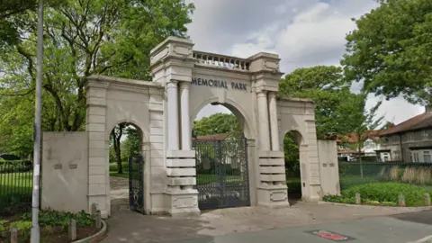 A trio of arches and columns forming the historic entrance to a park. The stone structure has the words 'memorial park' above the largest, central arch, with smaller arches on either side. There are metal gates within each arch.
