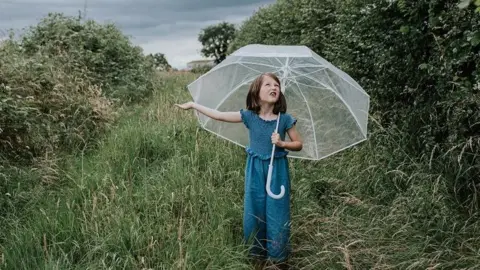 Getty Images Red-haired girl in blue denim holding a white umbrella with her arm out to check for rain and a questioning look on her face 