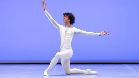 Gregory Batardon & Rodrigo Buas Jakob in a fully white dancers outfit with arms outstretched. He is on a stage, with a blue backdrop. He is smiling. He has short curly dark hair. 