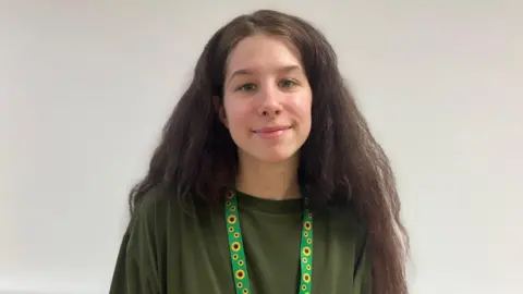 BBC Girl with green top and sunflower lanyard. She has long brown hair and a septum piercing and smiles at the camera.