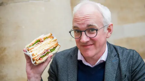 Emma Lynch/BBC The image shows a close-up of a person holding a large sandwich wrapped partially in white paper. The sandwich is thickly layered with sliced meat, melted cheese, leafy greens, and pieces of tomato between lightly toasted bread. The person is wearing round, dark-framed glasses along with a textured jacket over a collared shirt and a dark sweater. The background consists of a softly blurred, light-colored wall, which keeps the focus on the subject and the food.
