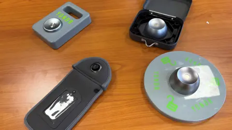 A black neoprene phone case sits on a brown wooden table. Three metal magnet removers sit around it. They have the word 'YONDR' printed on the front in green text.
