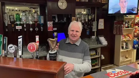 Doogie's Bar, Derrygonnelly John Doogan, a man with short, white hair, pulls a pint of Guinness behind his bar. He is wearing a striped grey jumper over a grey shirt. 