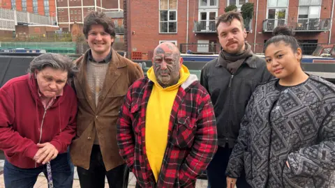 Four men and a woman in coats standing in a row in front of some large buildings