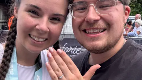 Sarah John holding up her hand with an silver engagement ring with a diamond. Sarah has brown plaits and is smiling. She is stood next to her partner Thomas, who has clear-framed glasses, a light beard and is smiling.