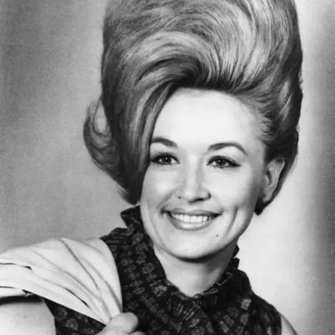 Getty Images//Michael Ochs Archives A black and white head shot of Dolly, smiling with a jacket slung over her shoulder. Her hair is nearly the same length as her face and stands back combed and high, with a curl leaning over to the left.