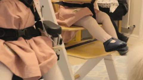 Two babies in pink outfits sat in highchairs next to each other. Picture shows their legs and the feet of one of them