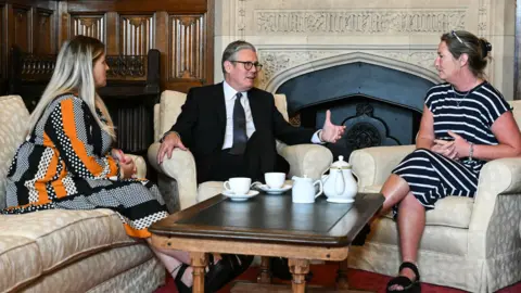PA Media Antonia Elverson (L) with a checked patterned blck and white with orange stripes is sitting on a sofa with the Prime Minister who has dark hair spectacles and a dark blue suit and her cousin Cheryl Korbel (R) who is wearing a black dress with thin white hoops