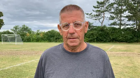 Ian Greaves, treasurer of Churchdown Panthers football club. He has grey hair and is wearing glasses and a grey jumper. He is standing on a football pitch with goalposts and bushes in the background.