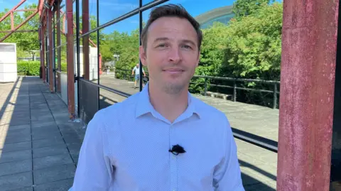 Sam Read/BBC Ryan looks at the camera wearing a white shirt with tiny black polka dots and a black buttonhole microphone. He is clean shaven and has short dark blonde hair. Behind him is one side of The Point, with reflective mirror panels showing a fence and trees, and blue sky.