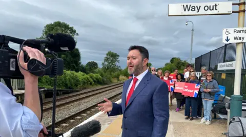 Sadik Al Hassan addressing the media on a train platform with a group of Labour supporters behind him