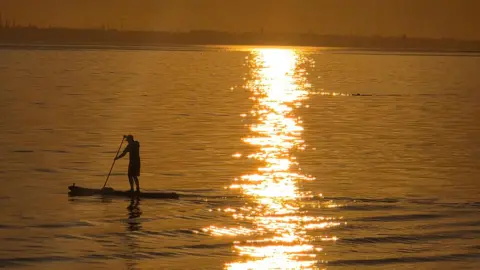 stacey johns The sun sets across the sea, with a paddleboarder silhouetted in the foreground.