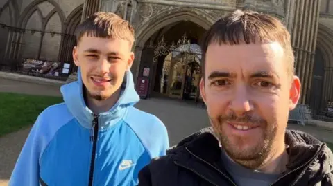 A photo of two men smiling in front of a building. Both have short brown hair and beards and the same haircut. The man at the front is wearing a black jacket and the man behind has a blue tracksuit on.