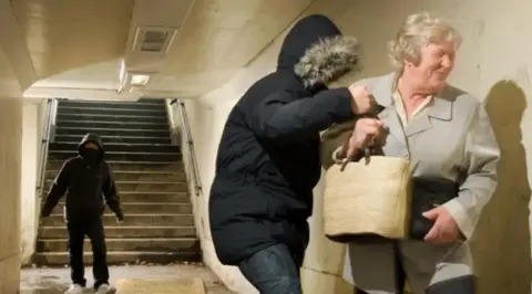 A woman in an underpass is manhandled by a youth who is taking her bag off her. Another youth stands a few feet away. The woman is grimacing. She is wearing a grey jacket and has short grey hair. The youths have their faces covered by furry-edged anoraks.