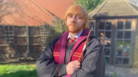 A man with ginger hair which is swept across his forehead and a ginger moustache and beard pulling a serious pose for the camera. He is wearing a Hogwarts school uniform with his arms crossed and a wand is in his hand.