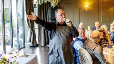 Gareth James A waiter in a black shirt and dark brown apron holds his hand out while singing into a microphone at a wedding. Guests can be seen sat on tables watching him sing.