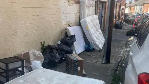 Bags of rubbish and mattresses dumped along a footpath on a residential street.