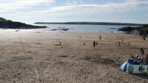 A sandy beach with people walking and a group in a wind breaker tent. the water and a peninsula are in the distance