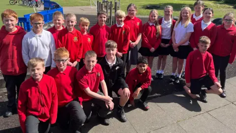 Boys and girls in red jumpers, red polo shirts and white shirts in two rows on a playground, looking at the camera.