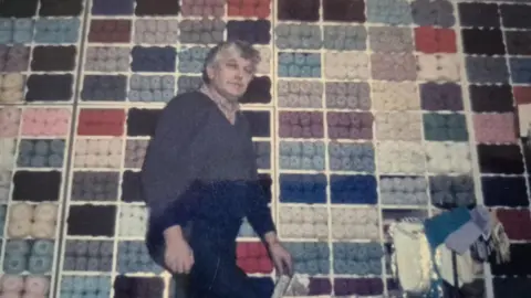 Christine's Wool Shop A man is wearing a long dark shirt and is standing on a ladder next to knitting yarns on shelves.