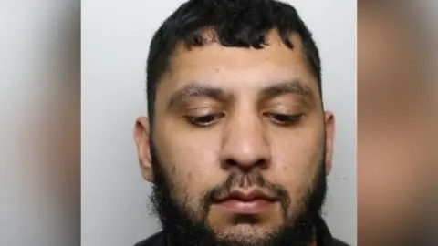 Police mugshot of Adam Iqbal. He has short black hair, brown eyes, a black beard and a thin black moustache.