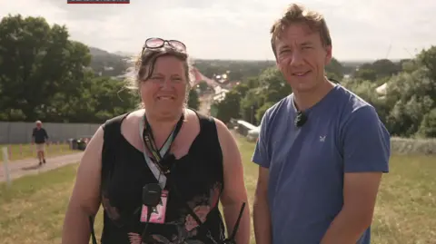Gareth Churcher and Jade Dunbar standing outdoors with a scenic background of trees and a distant view of a town or festival. Jade Dunbar wears sunglasses on their head, a black sleeveless top, and a lanyard. Gareth Churcher wears a blue t-shirt.