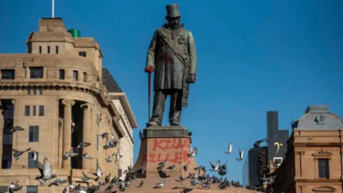 Gallo Images via Getty Images A statue of Paul Kruger in a square in Pretoria set against a cloudless blue sky. The word "killer" has been spray-painted onto the plinth below and pigeons are flying around the base of the statue.