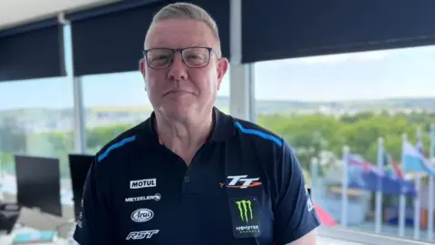 BBC Gary Thompson is wearing a TT polo shirt, wearing glasses, standing in front of large windows