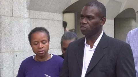 Daniel Anjorin's father delivers a statement outside court, he's wearing a black blazer and white shirt. Daniel's mother stands to the left of him, wearing a blue jumper and looking downwards.