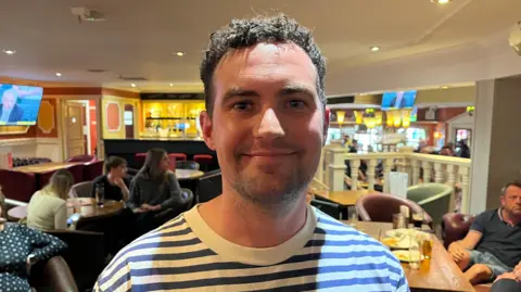 Oliver Conopo/BBC Josh Hendrickson smiling at Barratts wearing a stripy blue t-shirt