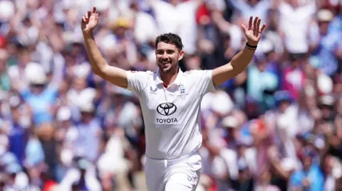 Josh Tongue wearing a white England cricket kit with the crest over his left breast and the Toyota logo on his stomach. He raises his arms in the air with a crowd in the background. 