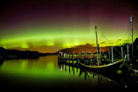 Owen Humphreys/PA Media The sky above the lake is illuminated orange, green and pink by the Northern Lights. Stars dot the night sky beyond it. The is a tall mountain in the background and a small boat pulled up to a wooden dock in the foreground.