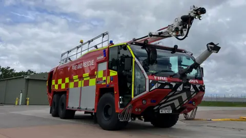 A large red and yellow fire engine is parked on an area of tarmac. It has two extendable arms - on its roof and one on the bonnet. Behind it is a distant airfield and a large grey hanger.