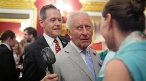 PA Vice Admiral Sir Tony Johnstone-Burt, Master of the Household to the Sovereign, wearing a black suit and red tie, holds a small battery powered fan for King Charles III, wearing a grey pinstripe suit and blue tie