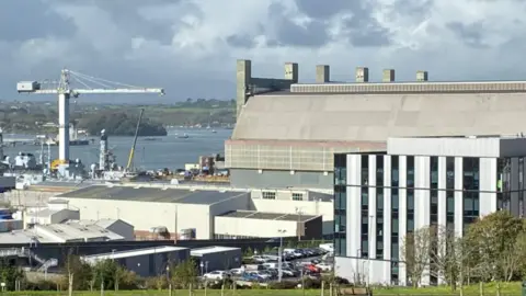 View of an industrial waterfront site with large warehouse buildings, a crane, and docked vessels beside a river or harbour. A modern office building and car park are in the foreground, with cloudy skies overhead.