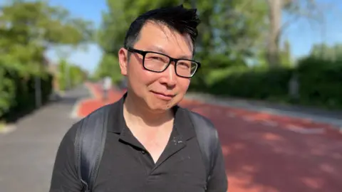 Tom Jackson/BBC Head and shoulders image of Will Yau. He has short black hair, dark trimmed glasses and is wearing a backpag and black polo shirt.