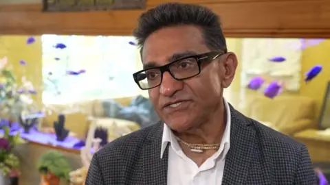 A man with glasses wearing a check jacket and a white shirt being interviwed for TV. 