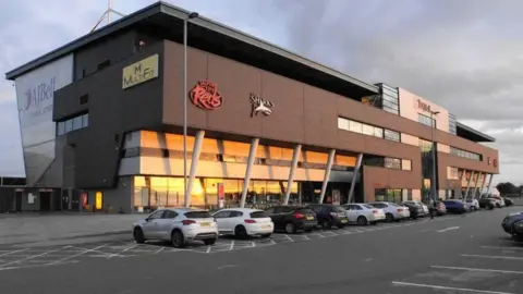 A view of the Salford Community Stadium from the stadium car park, with sunlight reflecting from the glass panelling along an arcade in front of the ground. 