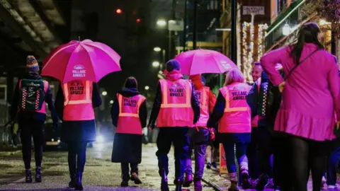 Village Angels A group of volunteers are walking along a street. They are wearing pink high-vis vests which read Village Angels 