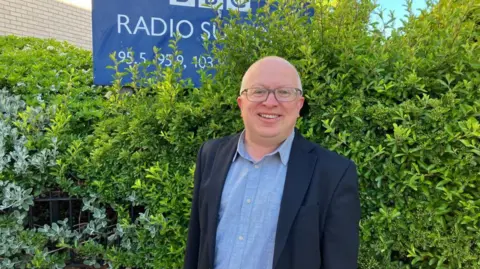 Vikki Irwin/BBC David Ellesmere is standing in front of the BBC Radio Suffolk building you can see a hedge to the right in the background and to the left there is a BBC Radio Suffolk sign. David is wearing a blue shirt and dark blue coat.