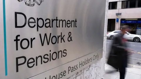 An out-of-focus person walks down a street with the sign for the Department for Work and Pensions in the foreground, outside Caxton House in London on 19 February