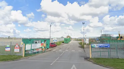 Google The entrance to Beverley household waste recycling site. Fencing surrounds the site. A car, a green shipping container and machinery can be seen in the distance. The sky is blue with white clouds. 