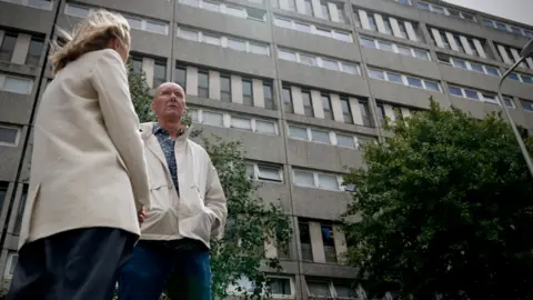 Adam Walker/BBC Katie Razzall with blond hair, white jacket and trousers talking to Irvine Welsh, bald, in white anorak and jeans outside the Banana flats in Leith