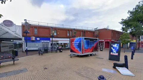 Google Maps General view of Bilston High Street including a row of red brick buildings with shops on the ground floor. There is a red and blue trailer in the centre of the picture.